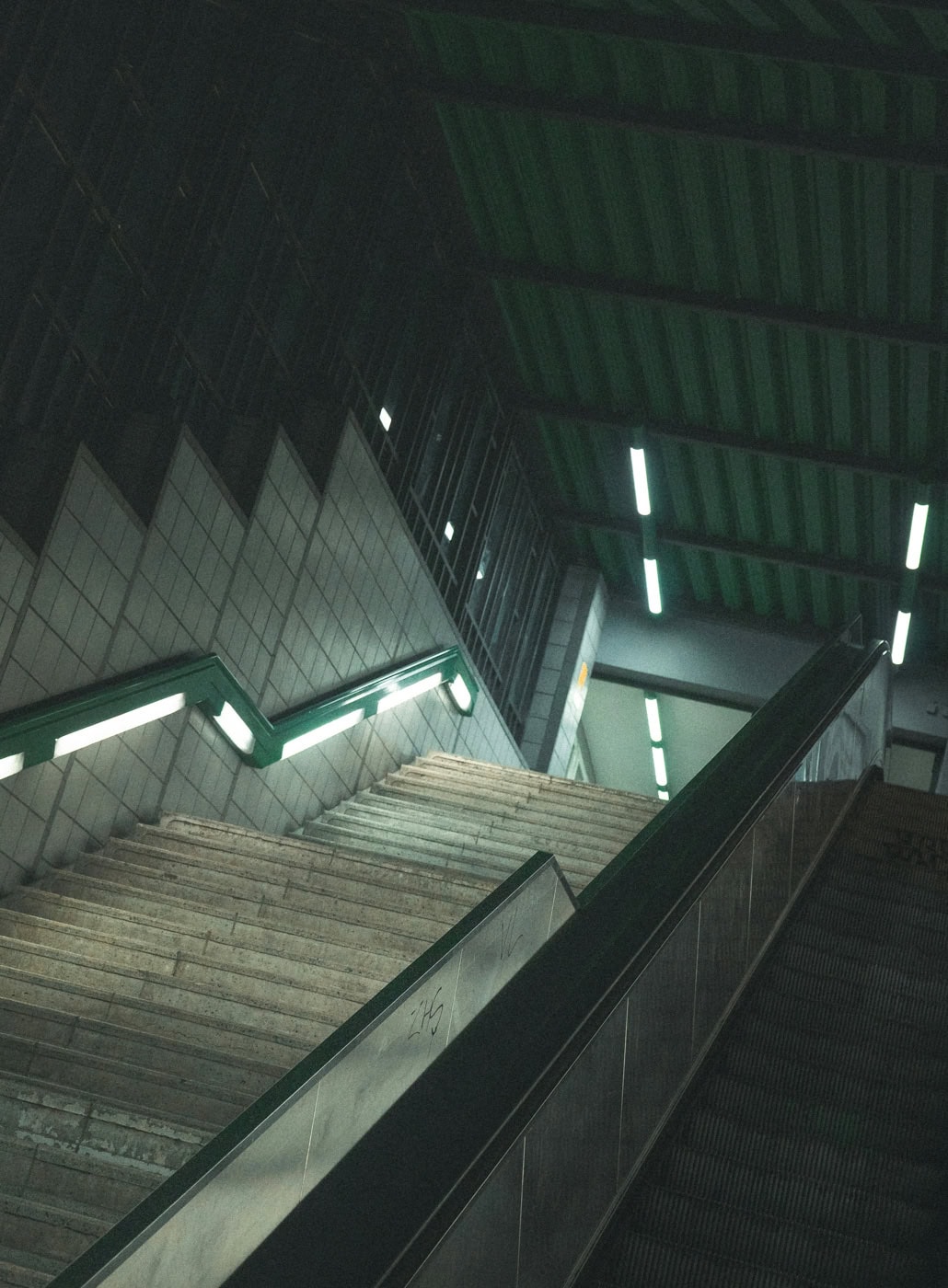 Ein U-Bahnhof mit einer steilen Treppe und einer Rolltreppe, beide führen nach oben. Die Szene ist in kühlem Licht gehalten.